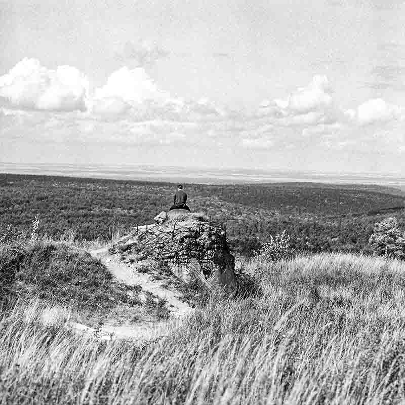 Historische Schwarz-Weiß-Aufnahme in der Normandie. Eine Person sitzt mit dem Rücken zur Kamera auf den Trümmern eines zerstörten Betonbunkers und blickt in eine weite, bewaldete Hügellandschaft unter einem leicht bewölkten Himmel. Im Vordergrund wächst hohes Gras.
