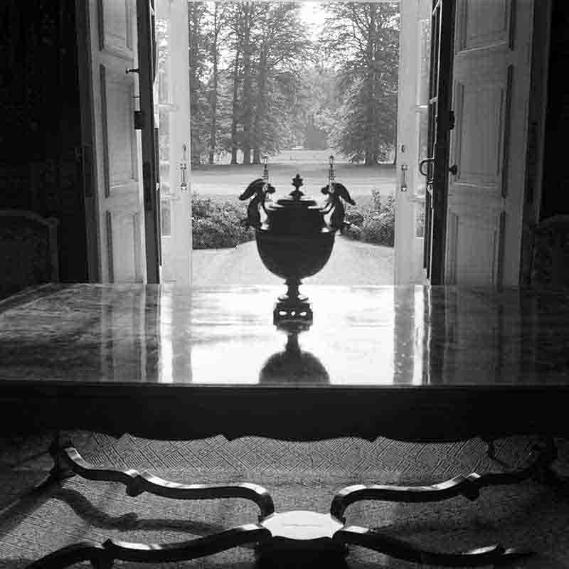Schwarz-Weiß-Fotografie von Peter Bock-Schroeder (1960) aus Haus Doorn: Ein prachtvoller Tisch im Vordergrund mit einer zentral platzierten, verzierten Prunkvase, die als Silhouette gegen das helle Licht weit geöffneter Flügeltüren kontrastiert.