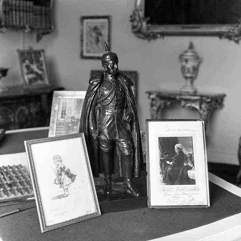 Historische Schwarz-Weiß-Aufnahme von Peter Bock-Schroeder (1960): Nahaufnahme des Schreibtisches von Kaiser Wilhelm II. im Haus Doorn. Im Zentrum steht eine Bronzestatue des Kaisers in Uniform mit Pickelhaube und Umhang, flankiert von persönlichen gerahmten Fotografien, darunter ein Porträt einer älteren Dame (Kaiserin Auguste Viktoria) mit handschriftlicher Widmung.