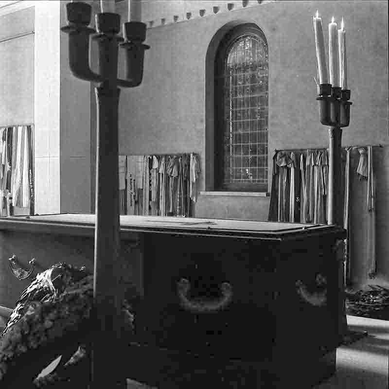 Schwarz-Weiß-Fotografie des Sarkophags von Kaiser Wilhelm II. im Mausoleum auf dem Gelände von Haus Doorn, aufgenommen von Peter Bock-Schroeder (1960). Der dunkle, massive Sarg steht im Zentrum, flankiert von hohen Kandelabern mit brennenden Kerzen. Im Hintergrund sind zahlreiche historische Fahnen und Standarten zu sehen, die an den Wänden hängen, sowie ein Rundbogenfenster mit Bleiverglasung.