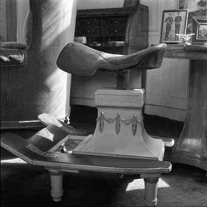 Detailaufnahme eines Reitschreibtisch-Geräts in Haus Doorn, fotografiert von Peter Bock-Schroeder (1960). Im Mittelpunkt steht ein auf einem verzierten, klassischen Säulenfuß montierter Ledersattel, davor eine klappbare, pultartige Ablagefläche mit Pedalen. Dieses mechanische Konstrukt ermöglichte es Kaiser Wilhelm II. im Exil in der gewohnten Haltung eines Reiters zu arbeiten